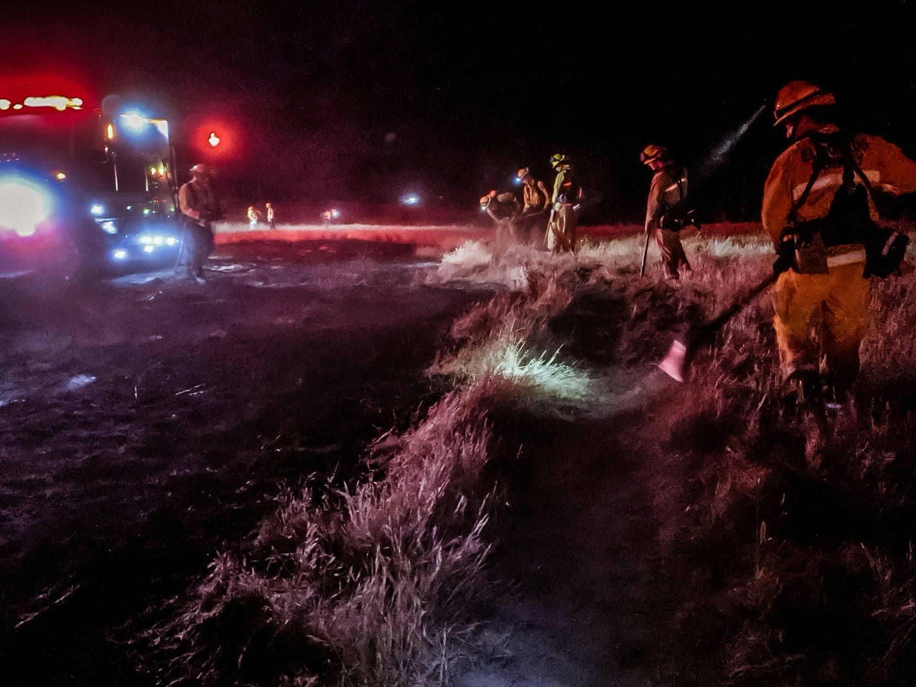 Firefighters are battling a fire at night, equipped with flashlights and tools, with emergency vehicles and red lights nearby.