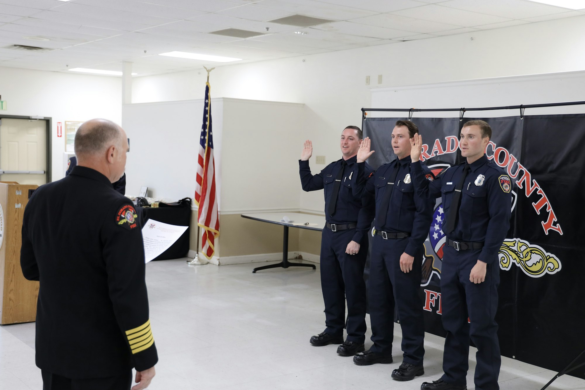 Firefighters taking an oath during a ceremony with an official presiding, American flag in the background.