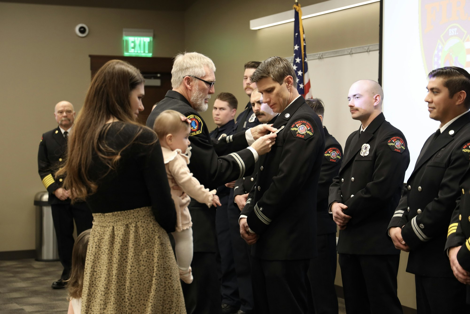 A firefighter is being pinned with a badge in a ceremony, surrounded by other uniformed individuals and family members, including a baby.