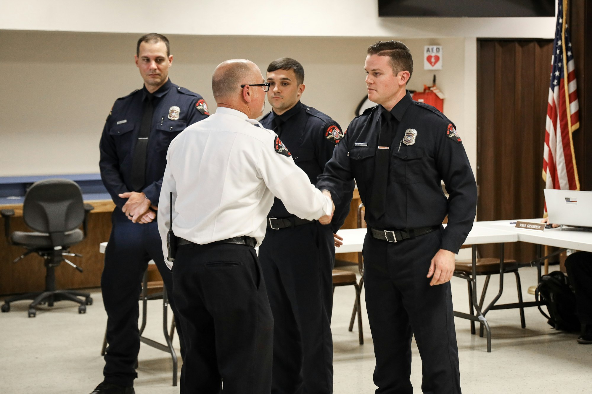 Four firefighters in uniform, one shaking hands with another, inside a room with an American flag.