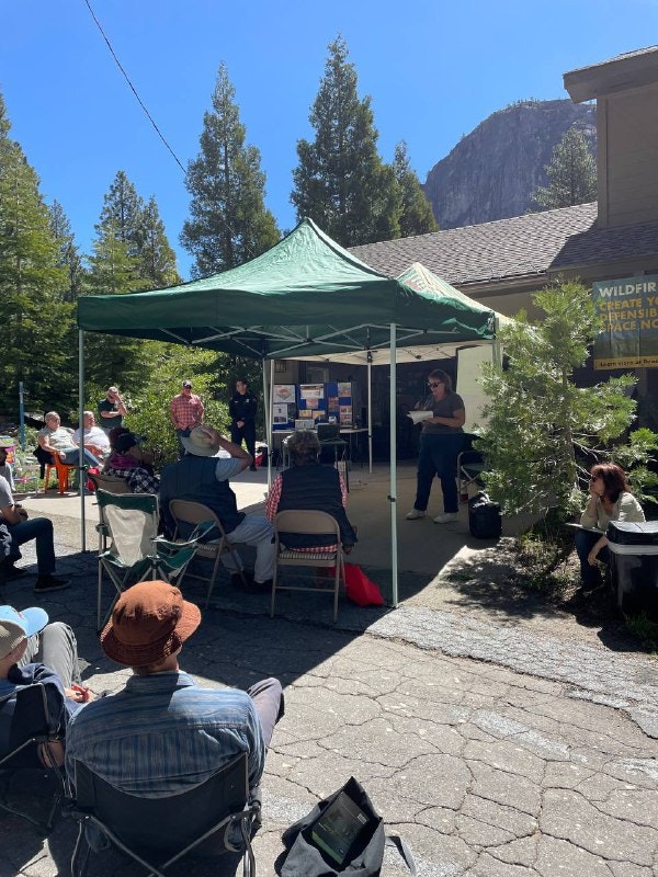 A group attending a presentation under a tent in a forested area, discussing wildfire preparedness.