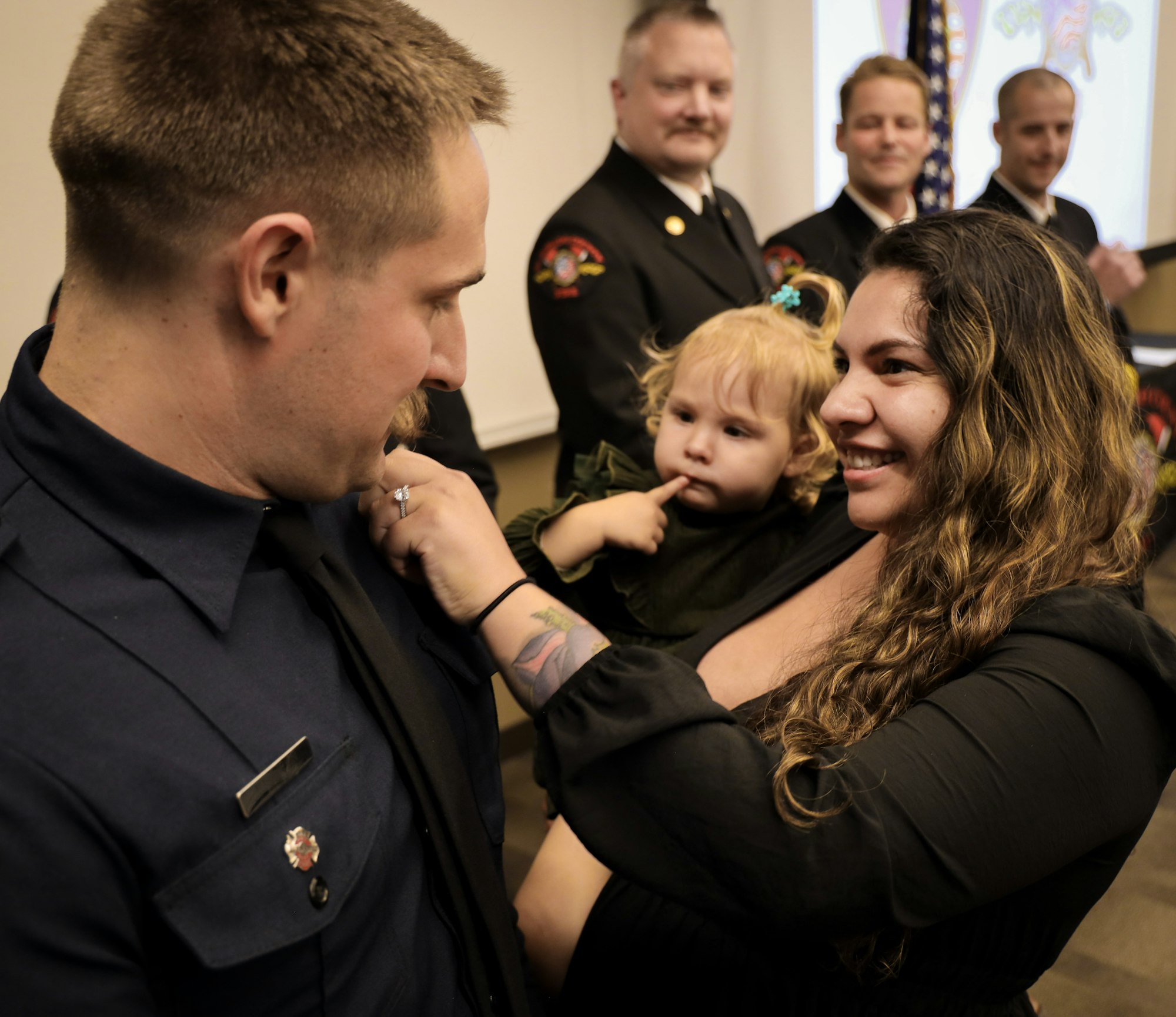 A firefighter is smiling and interacting with his partner and their young child during a celebratory moment in an official setting.
