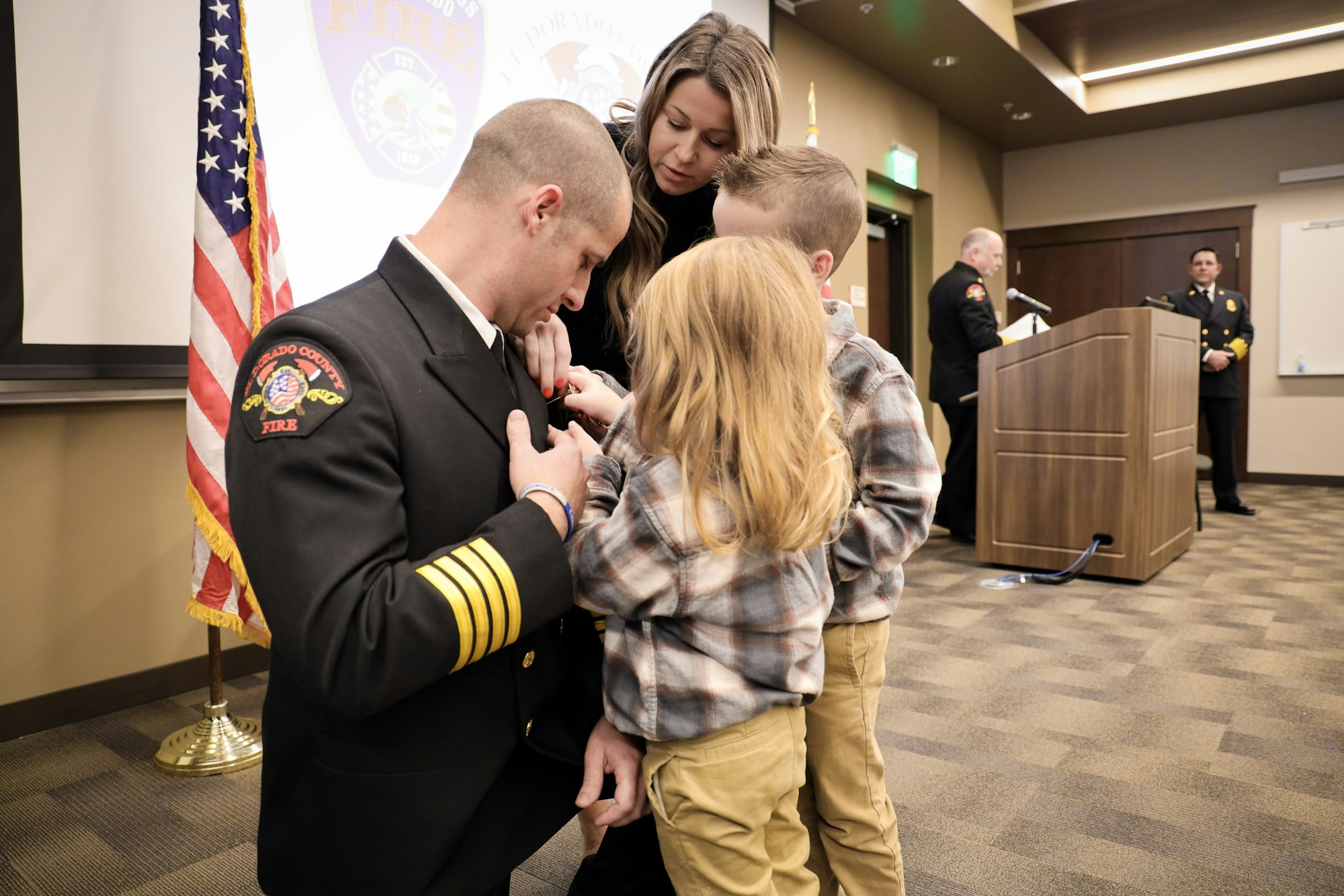 A firefighter is being honored with family present, as children and a woman assist with his uniform during a ceremony.
