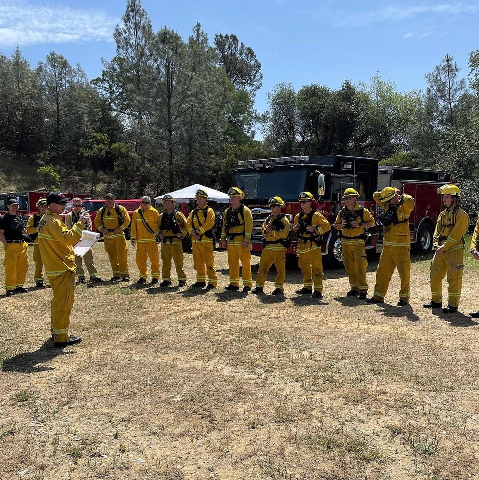 A group of firefighters in yellow gear is gathered for a briefing near a fire truck in a wooded area on a sunny day.