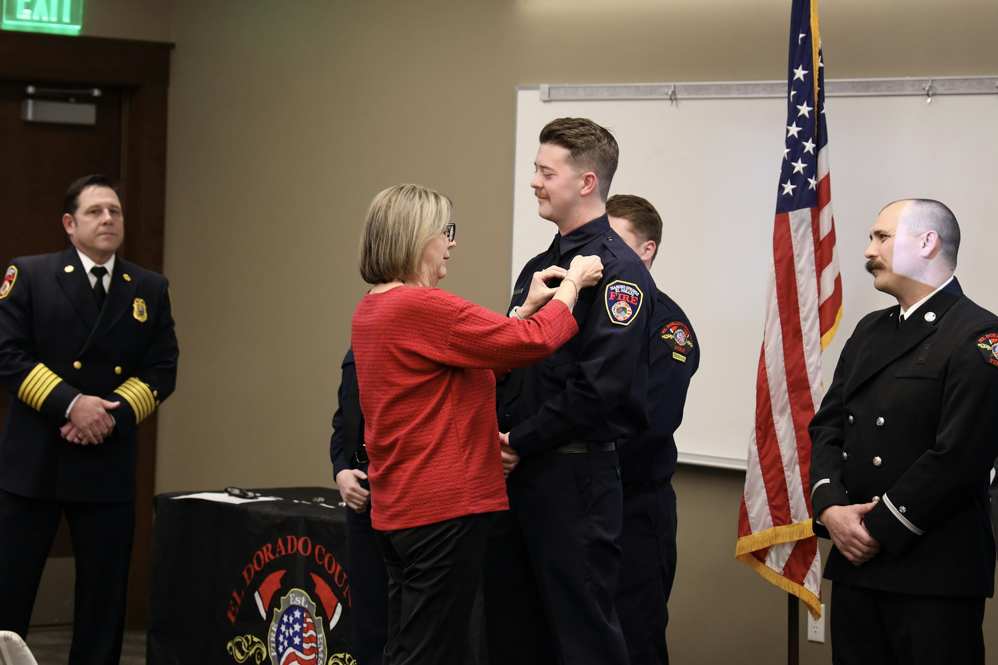 A woman pins a badge on a firefighter in uniform during a ceremony. Other uniformed personnel stand by. An American flag is displayed.