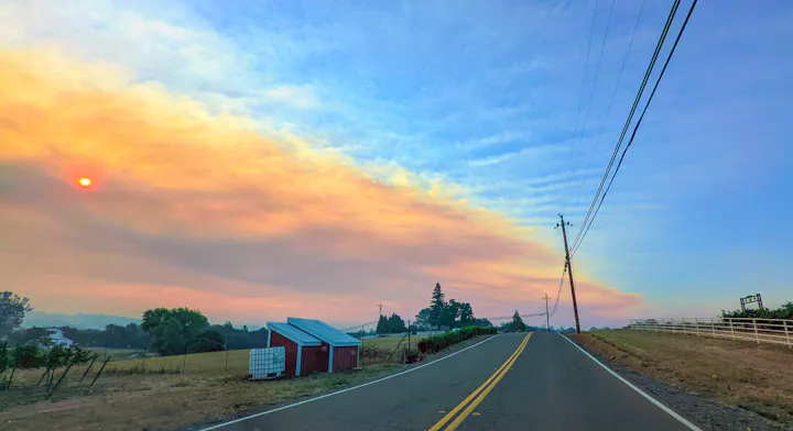 A rural road with a smoky sky, a sun partially obscured by haze, utility poles, and a small building on the side.