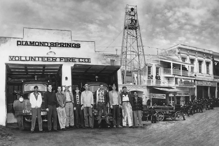 A historical photo of volunteer firemen posing outside their station in Diamond Springs, with fire trucks and a tall lookout tower.
