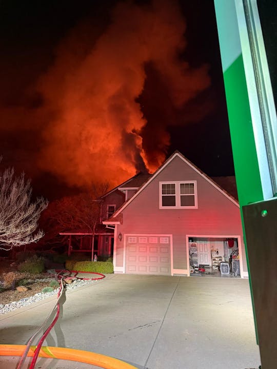 A house on fire at night with smoke billowing, hoses on driveway, and open garage door.