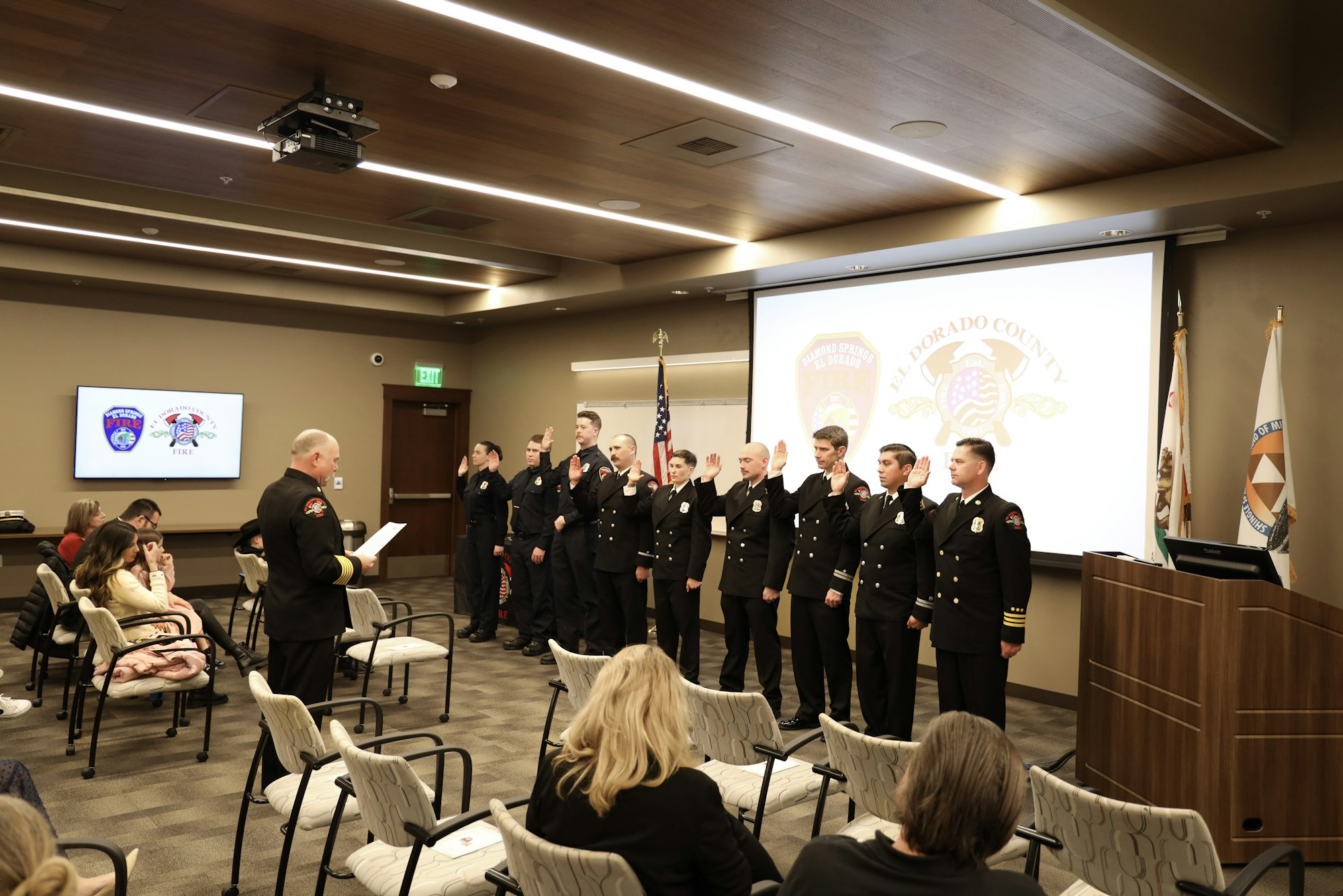 A group of uniformed individuals stands in formation for a ceremony in a room, with others seated watching. Flags and a screen are visible.
