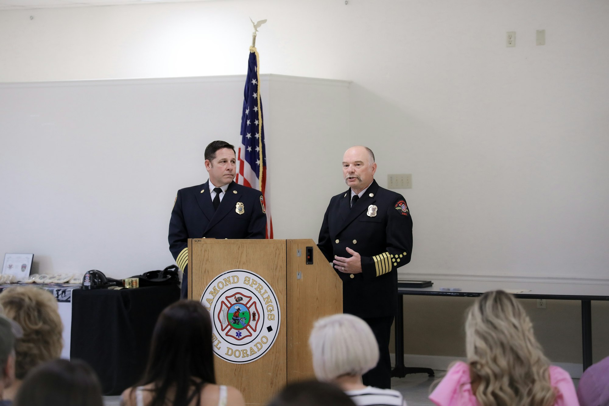 Two firefighters at a podium, audience listening, U.S. flag in the background.