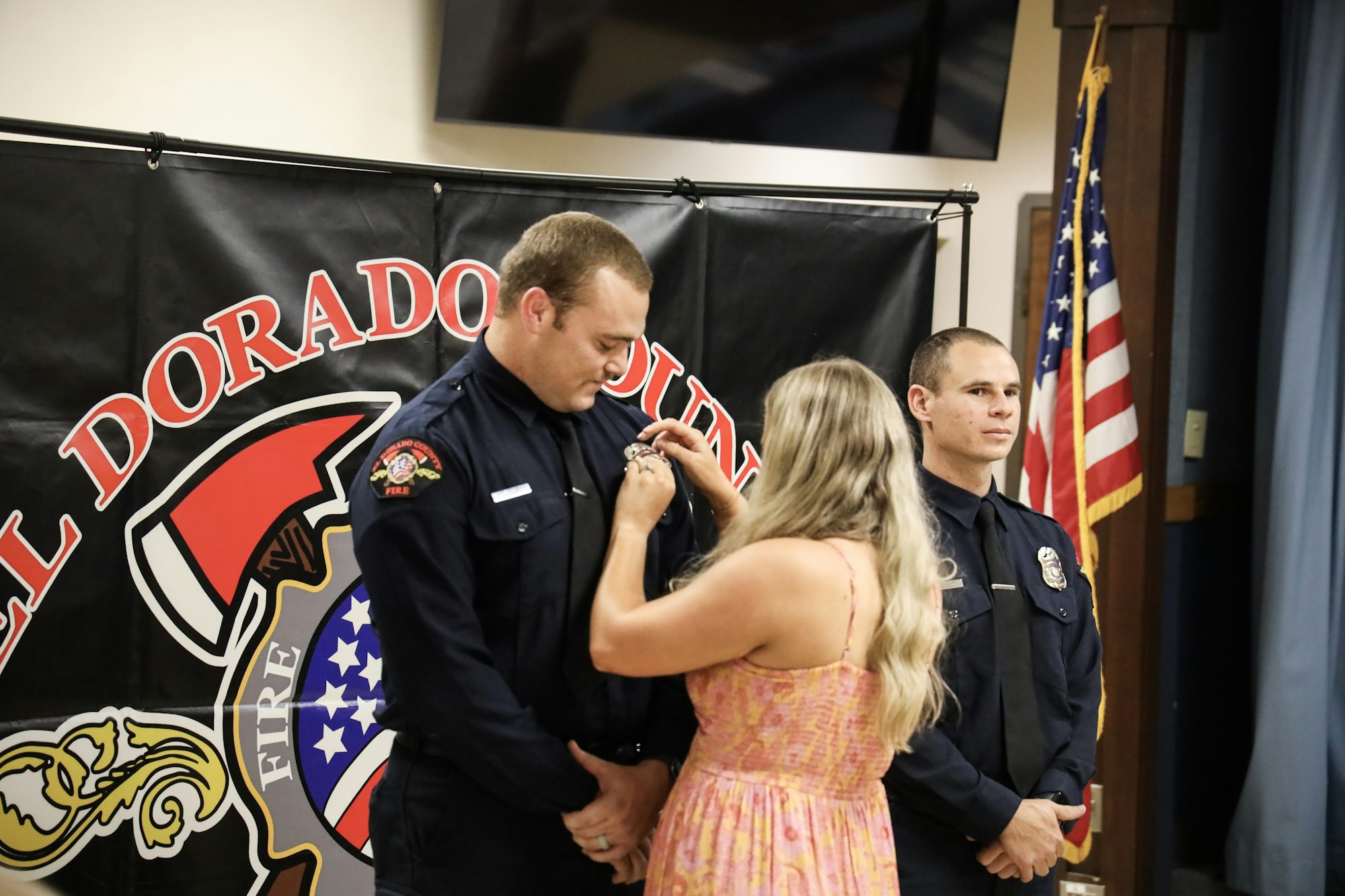 Woman pinning badge on firefighter's uniform, another firefighter observing, American flag in background, "El Dorado County Fire" banner.