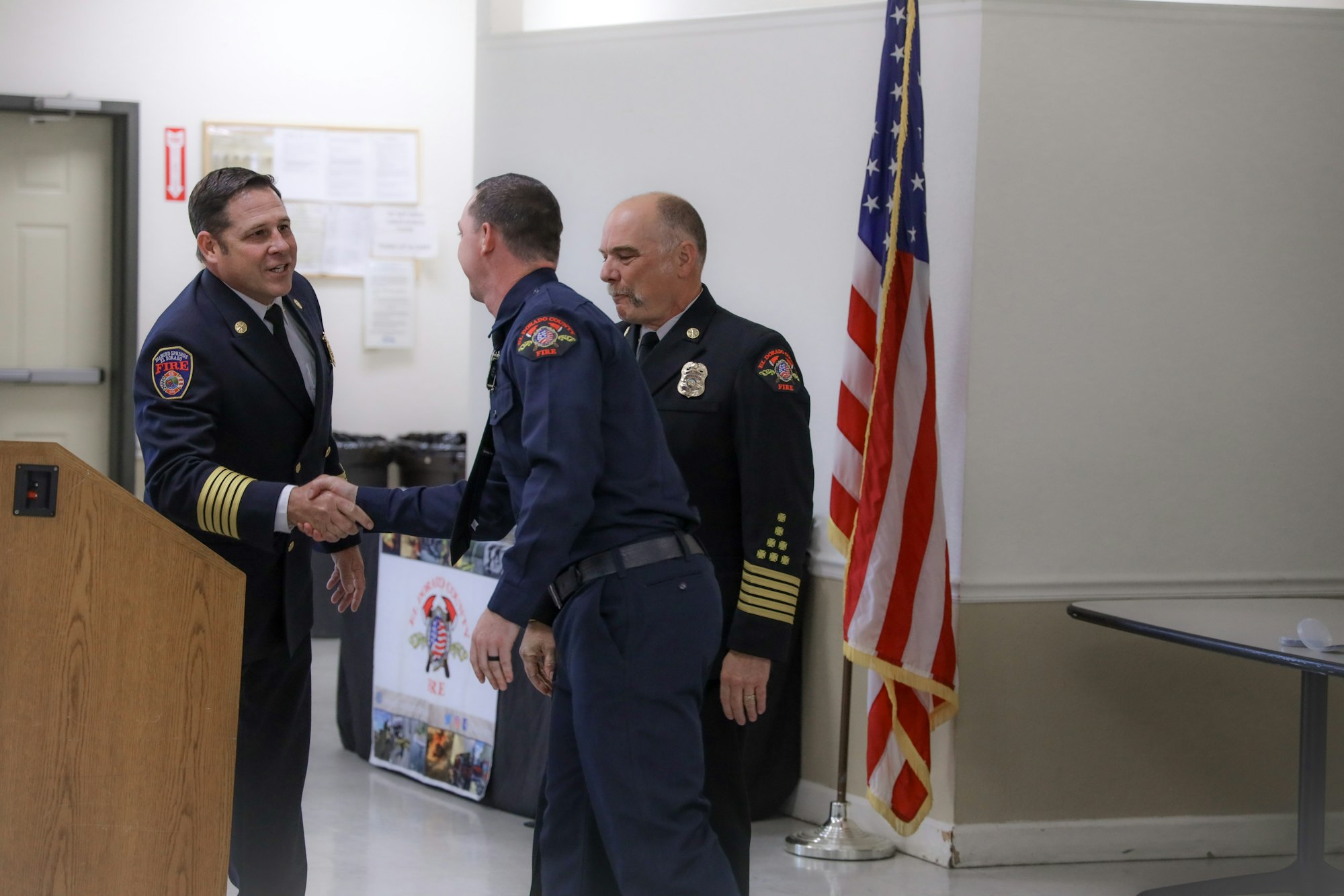 Three firefighters in uniform shaking hands in a room with an American flag.