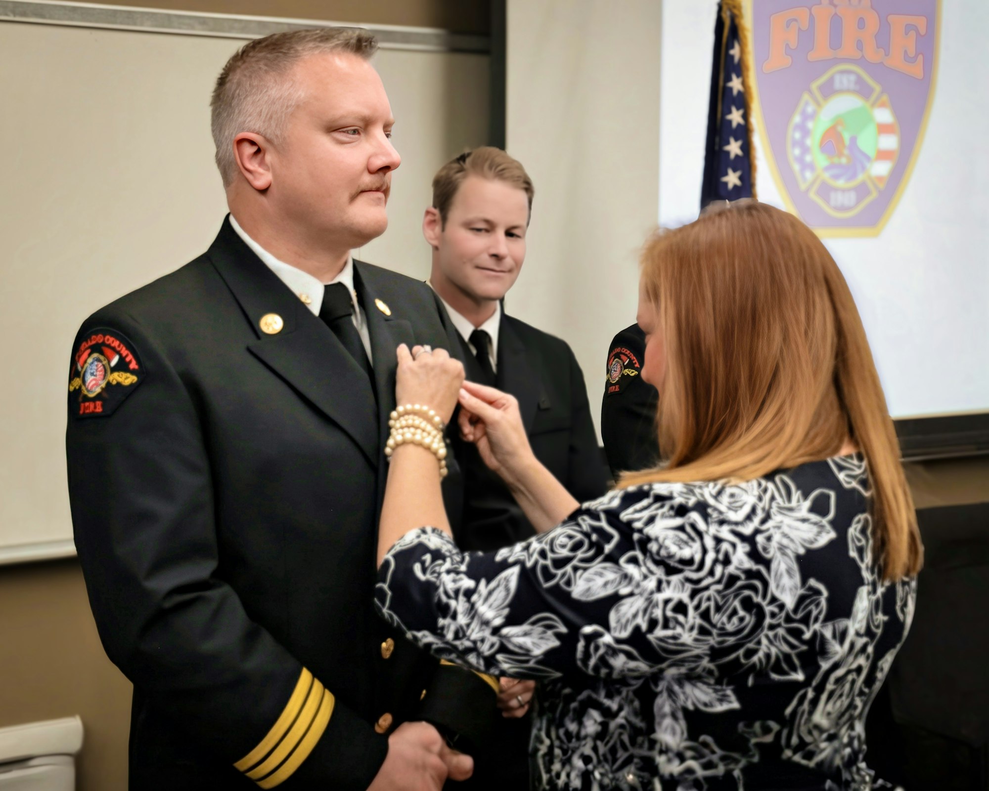A ceremony where a woman pins a badge on a firefighter, honorably recognizing his service, with another firefighter in the background.