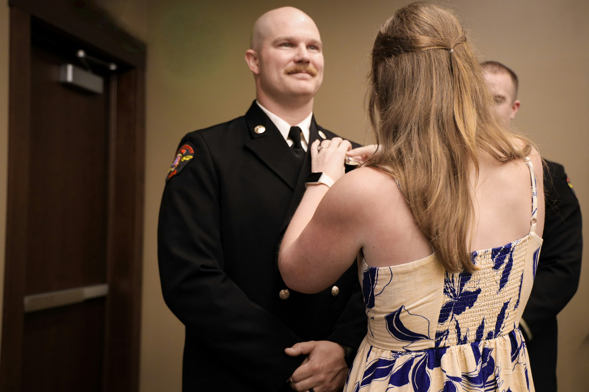 A woman in a dress helps a man in a fire uniform with a ceremonial pin or badge, celebrating a special moment together.