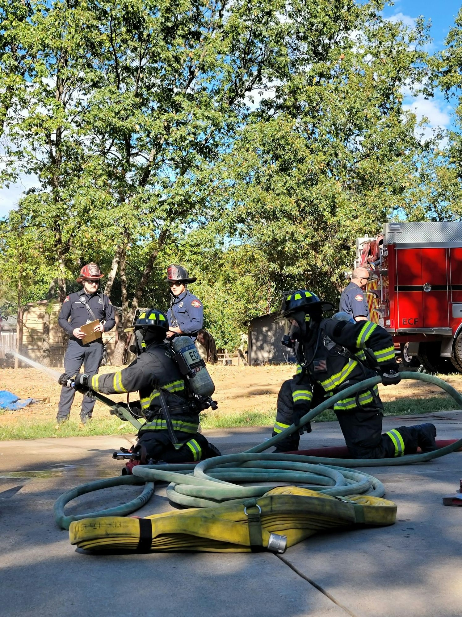 Firefighters in gear practice with hoses, while two officers observe and a fire truck is parked nearby. Trees and a blue sky are visible.