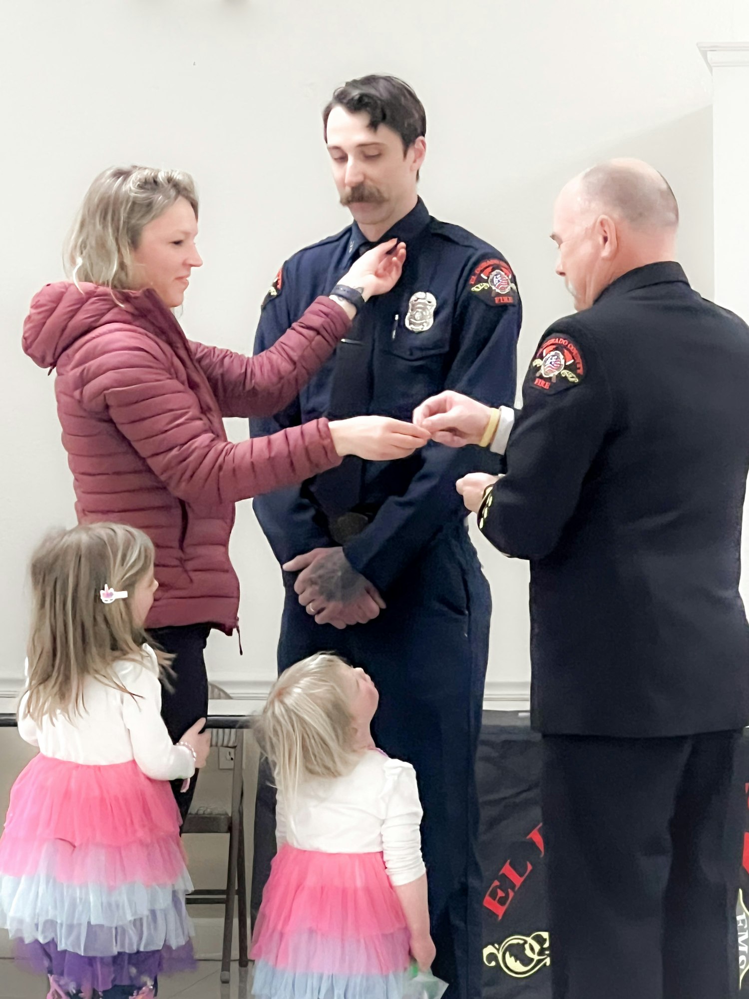 A woman pins a badge on a firefighter's uniform, two children watch, another firefighter assists, in an indoor ceremony.