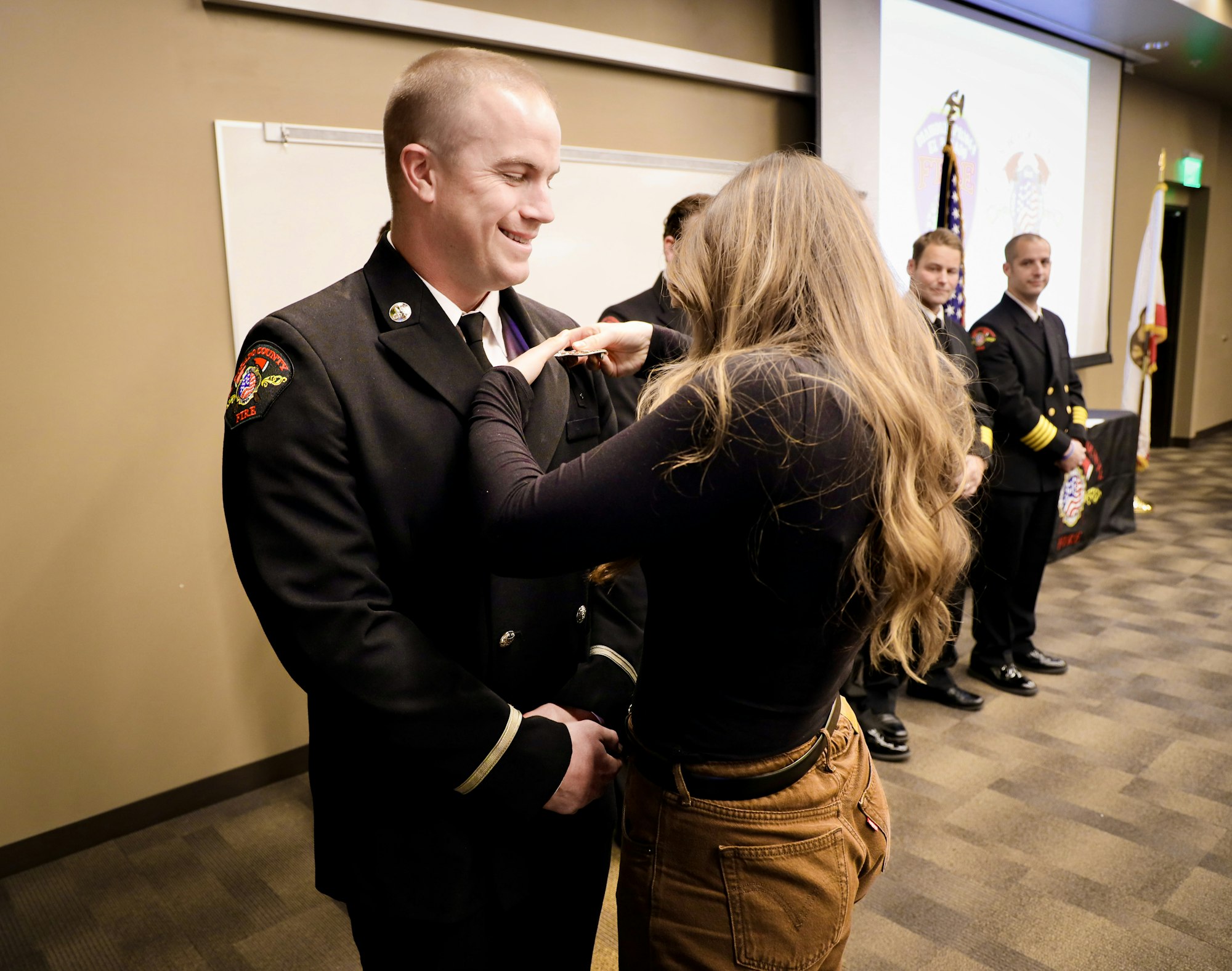 A firefighter is receiving a badge from a woman in a formal ceremony, with others observing in the background.