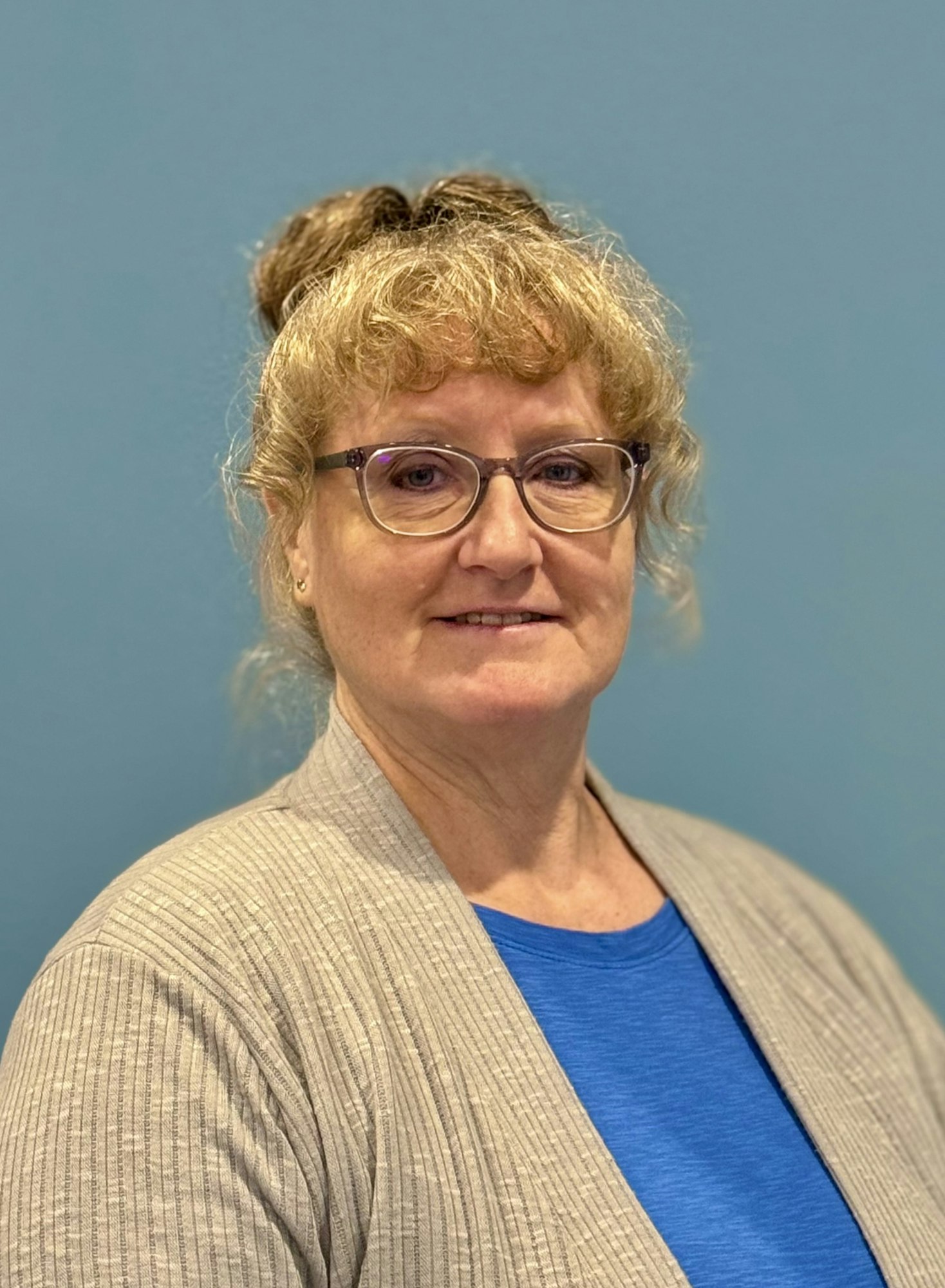 A woman with glasses, a topknot hairstyle, a blue top, and a beige cardigan against a blue background.