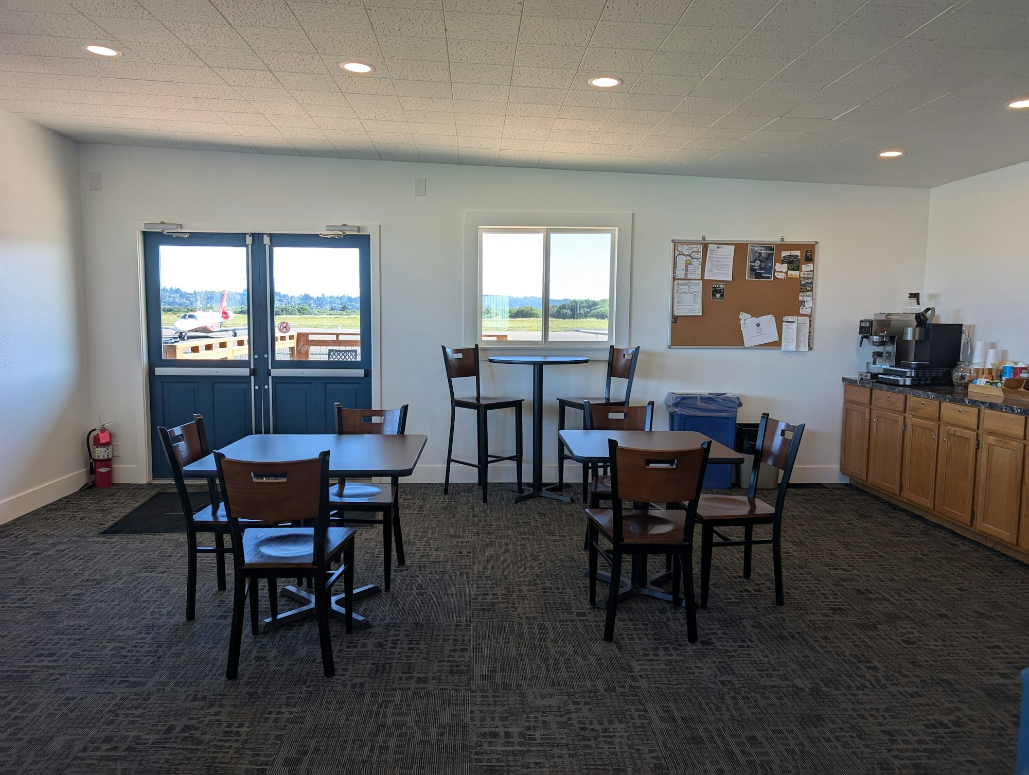 A small room with tables, chairs, notice board, coffee machine, blue doors, and a window overlooking a landscape.