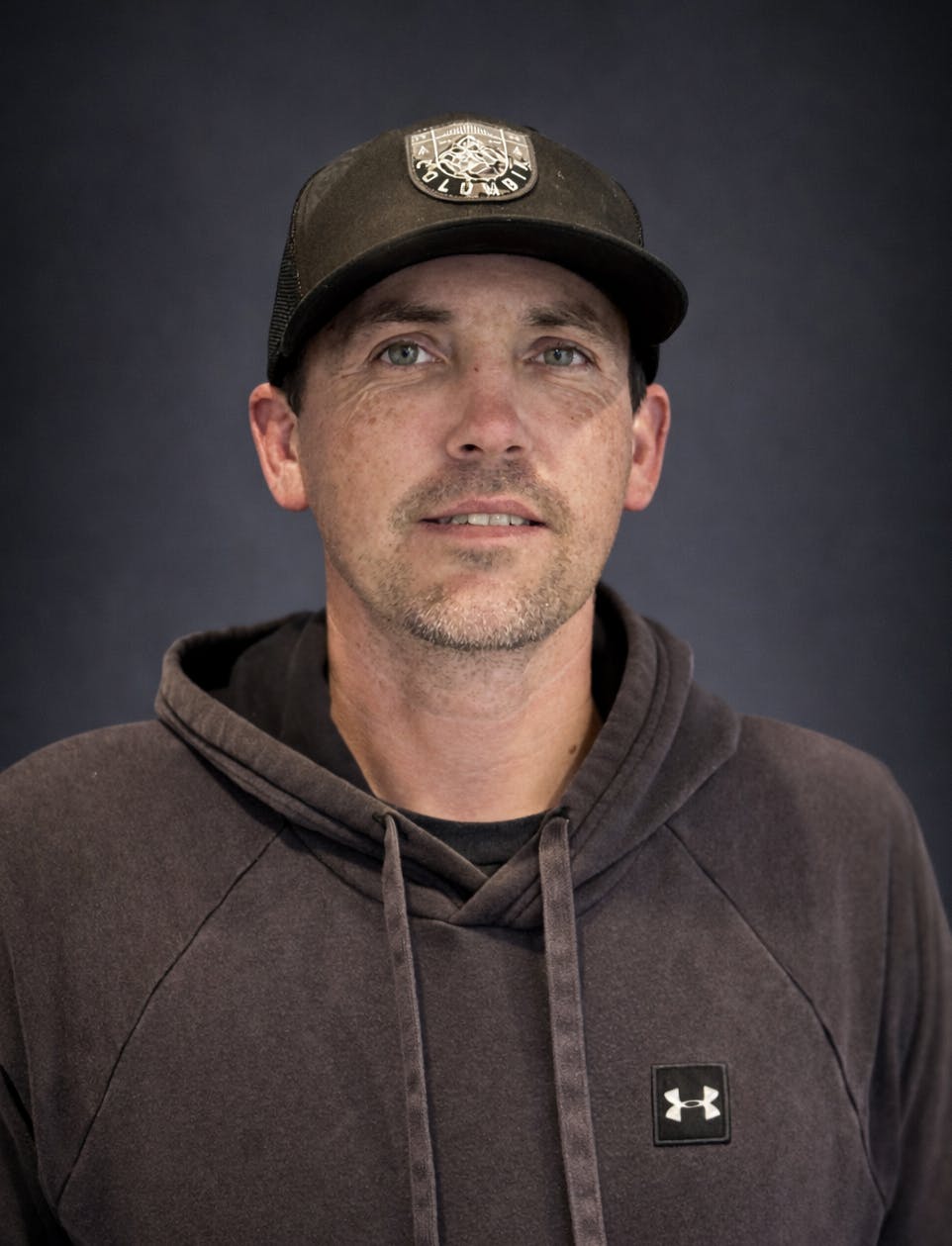 A man is wearing a hoodie and a baseball cap, posing against a dark background. He has a relaxed expression.
