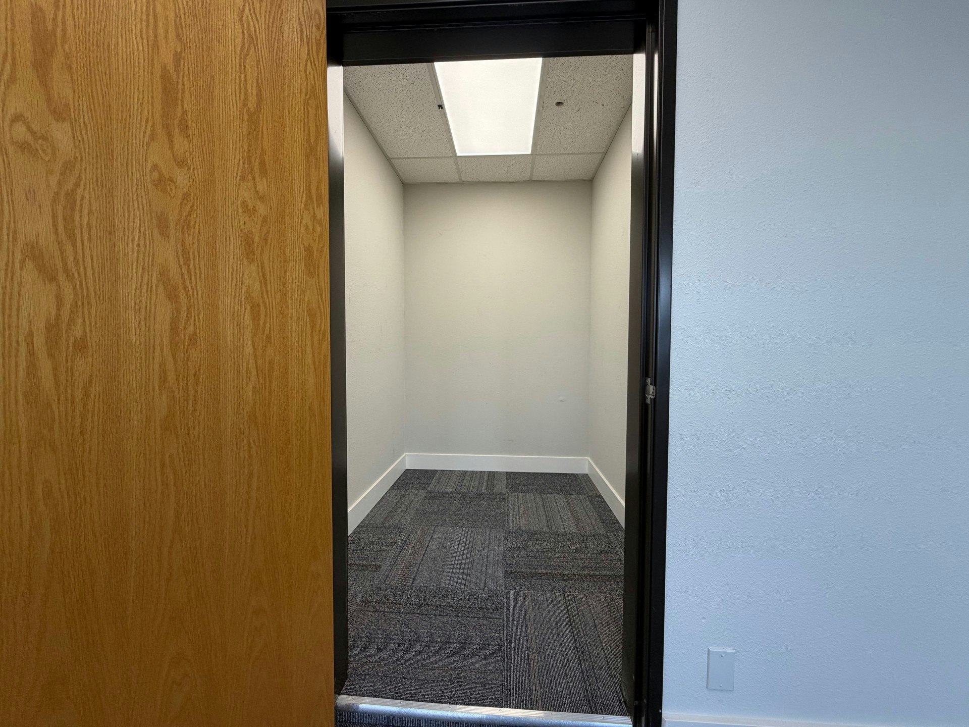 The image shows an open door leading into a small, empty office space with gray carpet and plain white walls.