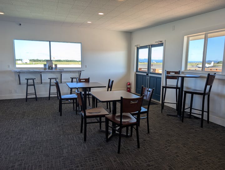 Room with tables and chairs, large windows showing an outdoor airport scene with planes, and a fire extinguisher by the doors.