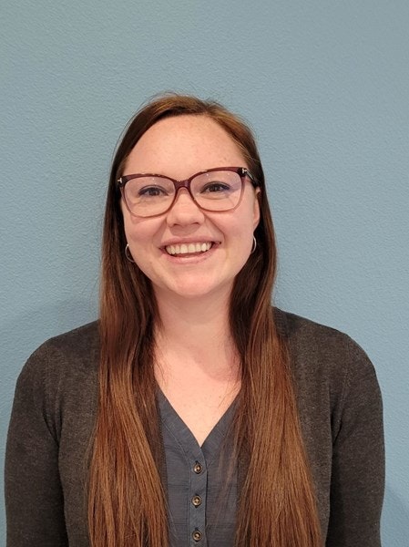 A smiling person with glasses, long hair, and a cardigan against a blue wall.