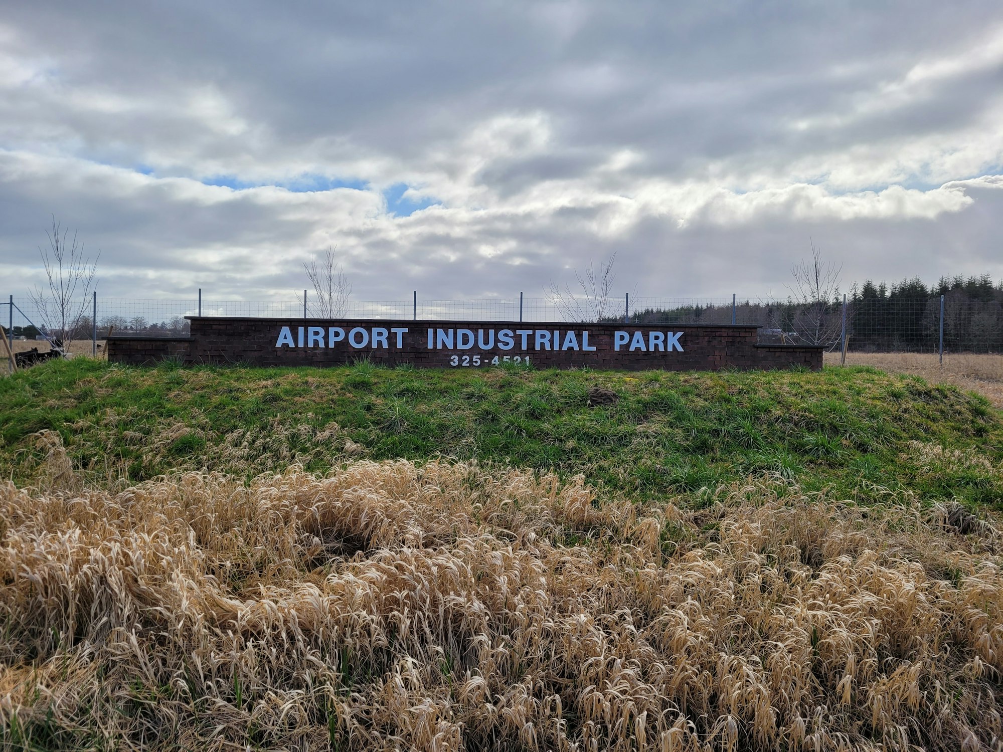 Sign reads "Airport Industrial Park" with phone number, set in a grassy field under a cloudy sky.