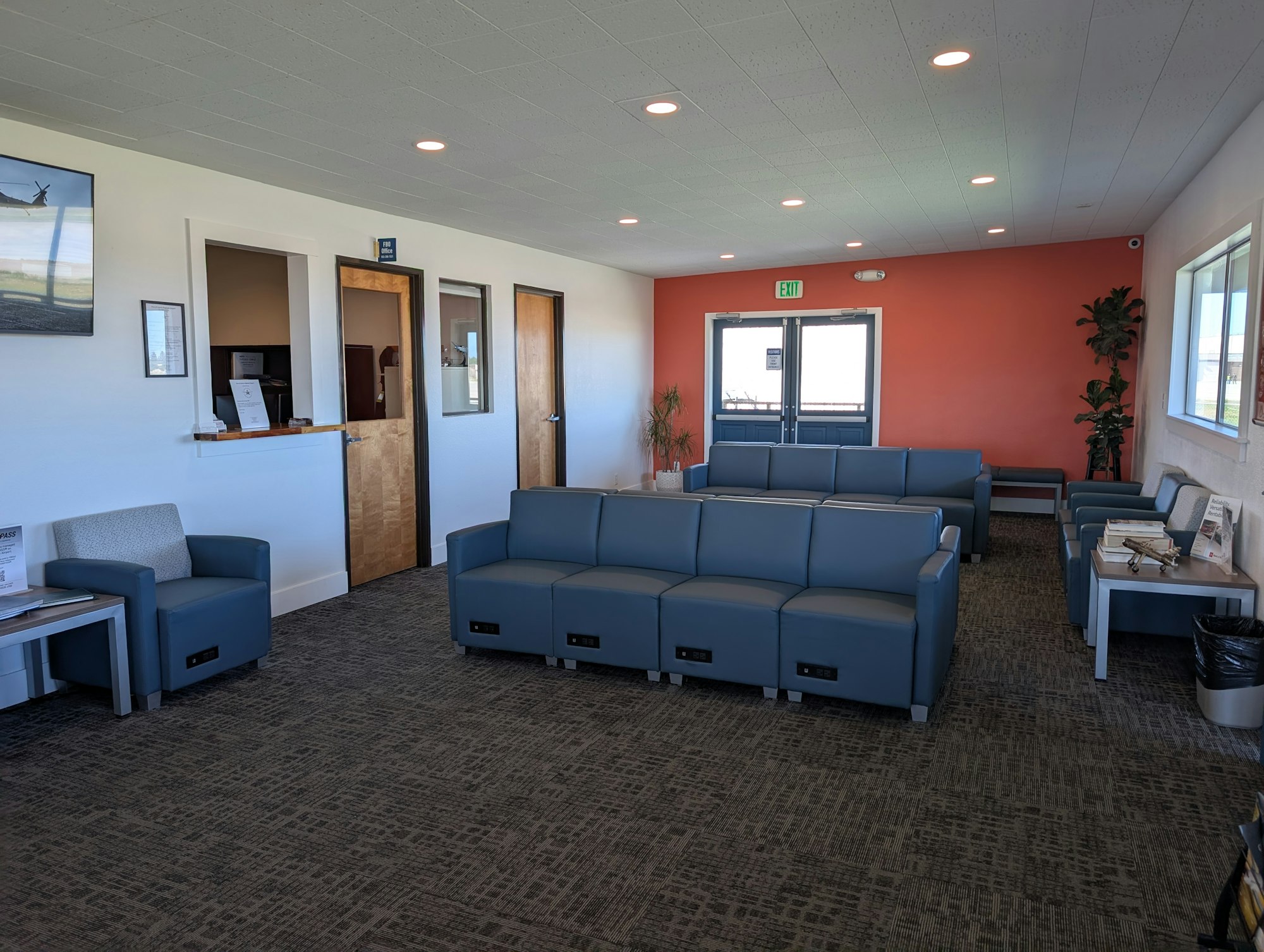 A waiting room with blue seats, a red wall, plants, and magazines on tables.