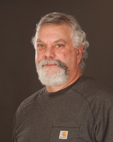 A man with grey hair and beard, wearing a grey t-shirt, against a dark background.