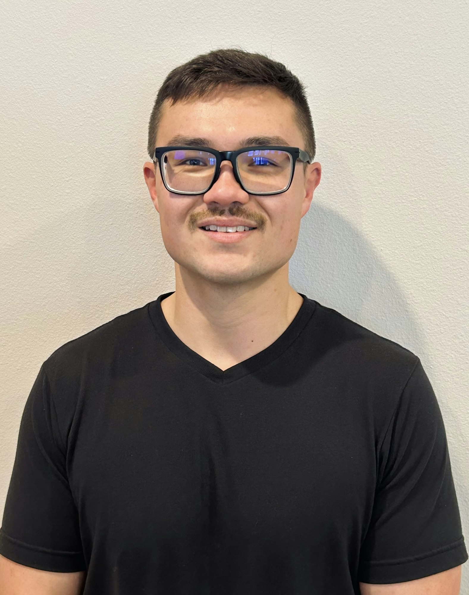 A person wearing glasses and a black shirt, smiling against a plain wall.