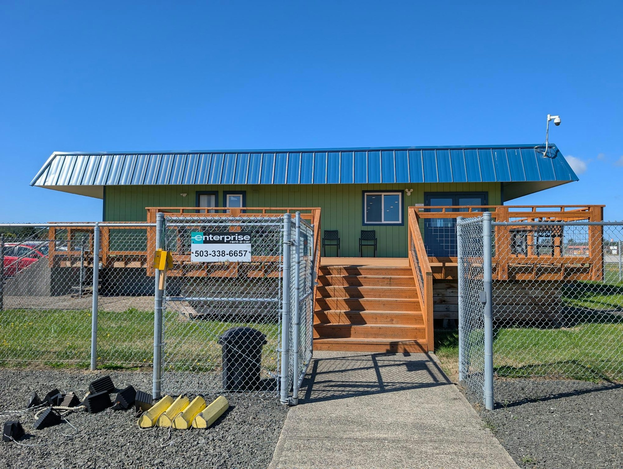 A fenced building with a blue roof, stairs, and a sign with a phone number on the gate, under a clear blue sky.