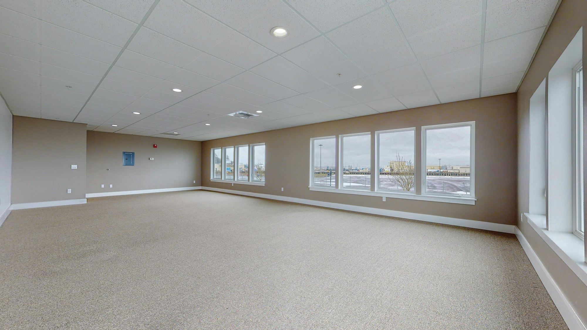 The image shows a spacious, empty room with carpeted flooring, ceiling lights, and large windows.