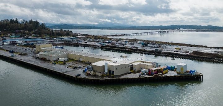 Industrial buildings on a waterfront with a pier and distant hills under cloudy skies.
