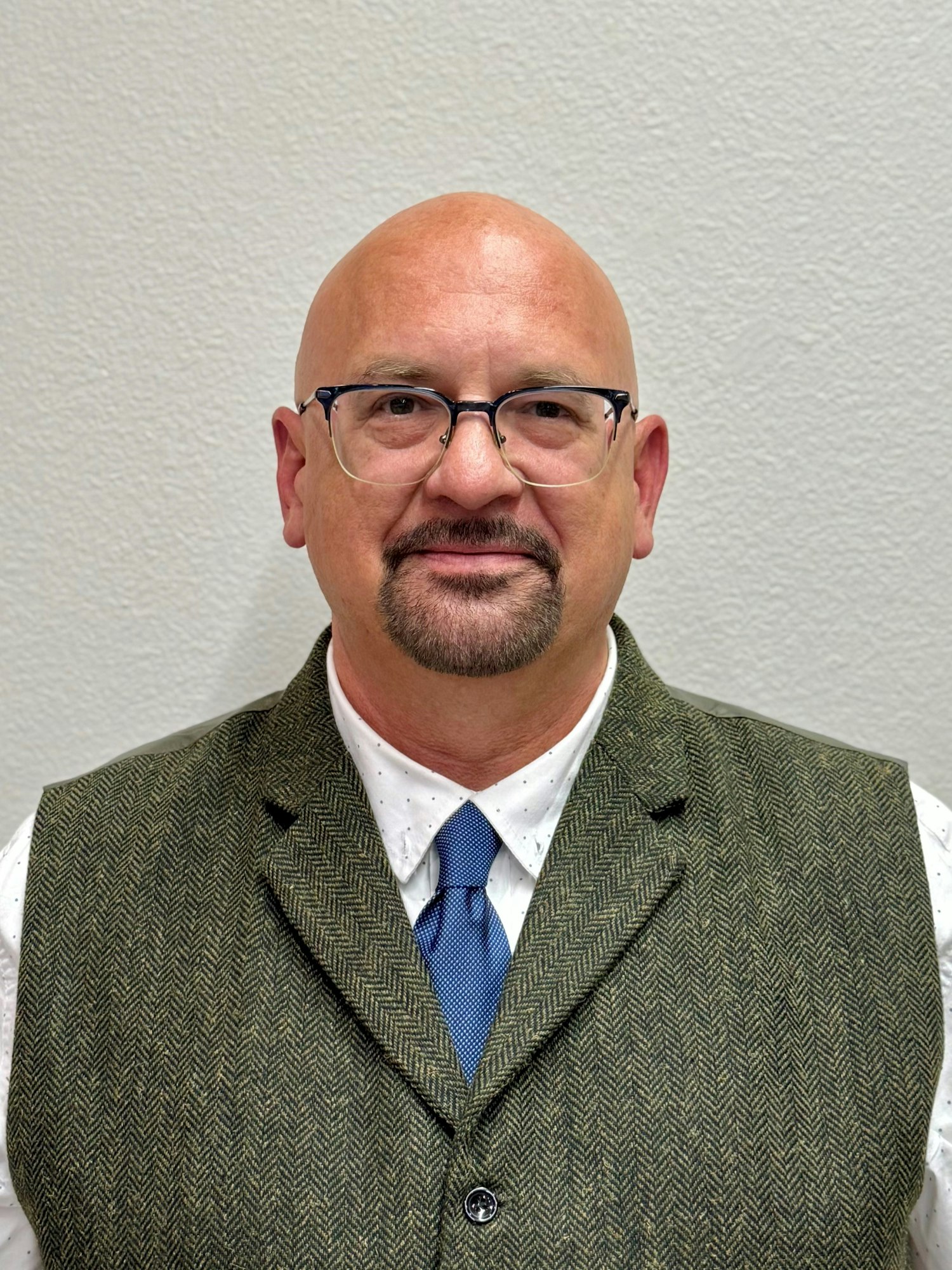A person wearing glasses, a herringbone vest, white shirt, and blue tie, standing against a light-colored wall.