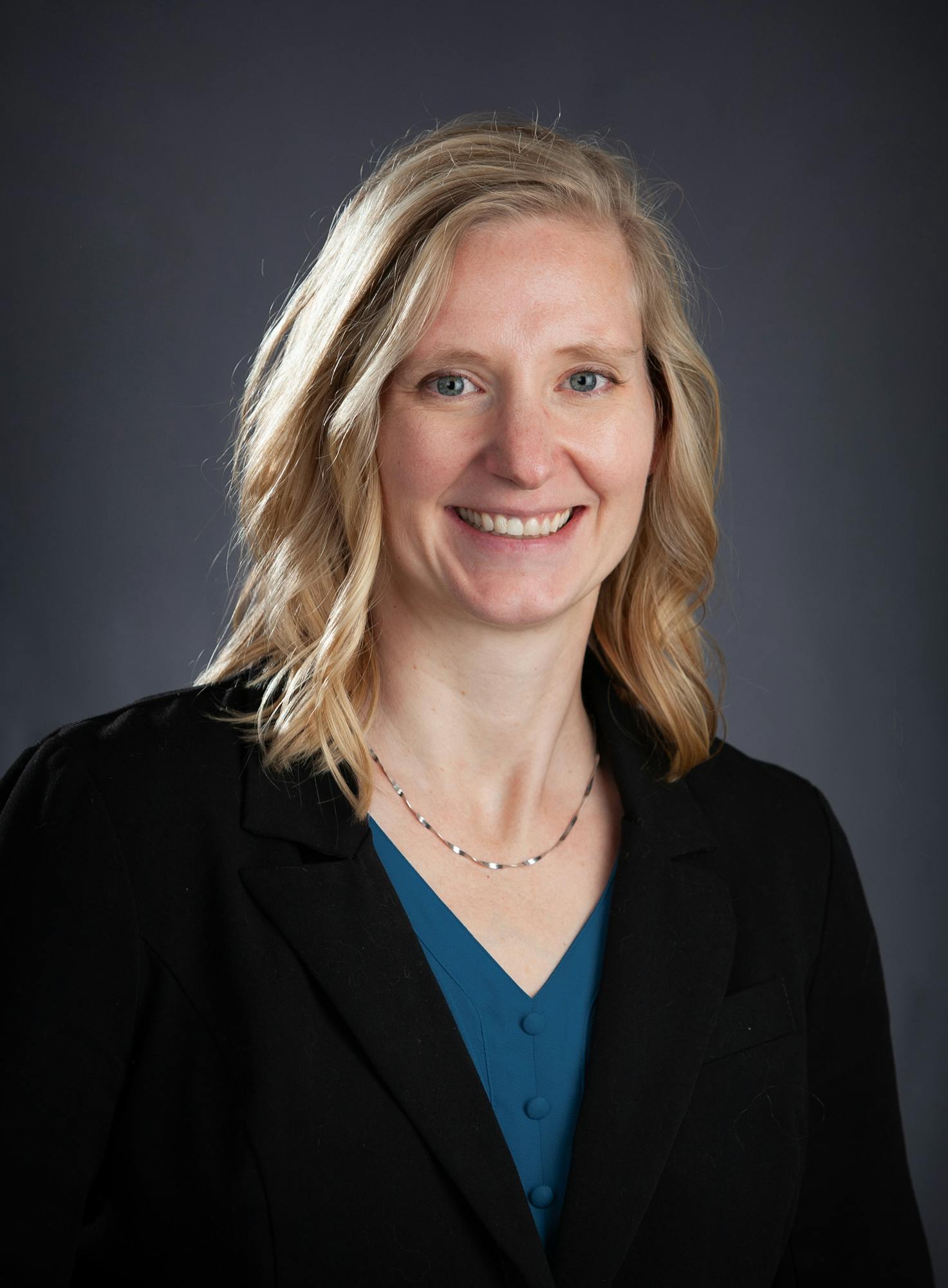 A woman with shoulder-length blonde hair smiles in a black blazer over a teal top, against a dark background.