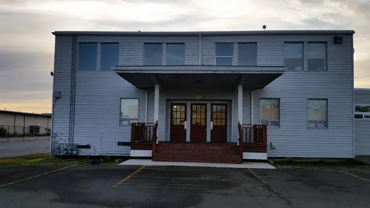A two-story building with a wooden entrance and steps, featuring multiple windows and a cloudy sky in the background.