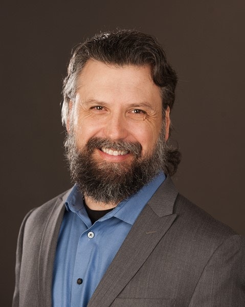 A smiling man with a beard, wearing a blazer over a blue shirt, against a dark backdrop.
