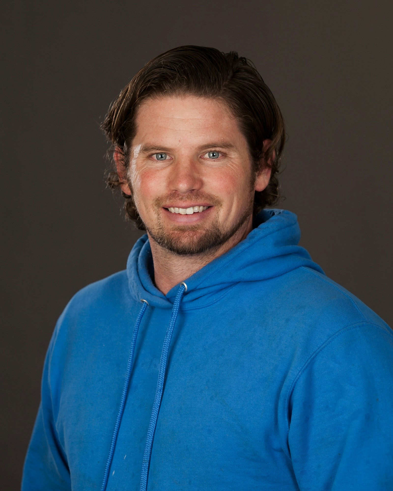 A smiling man with medium-length hair wearing a blue hoodie, against a gray background.