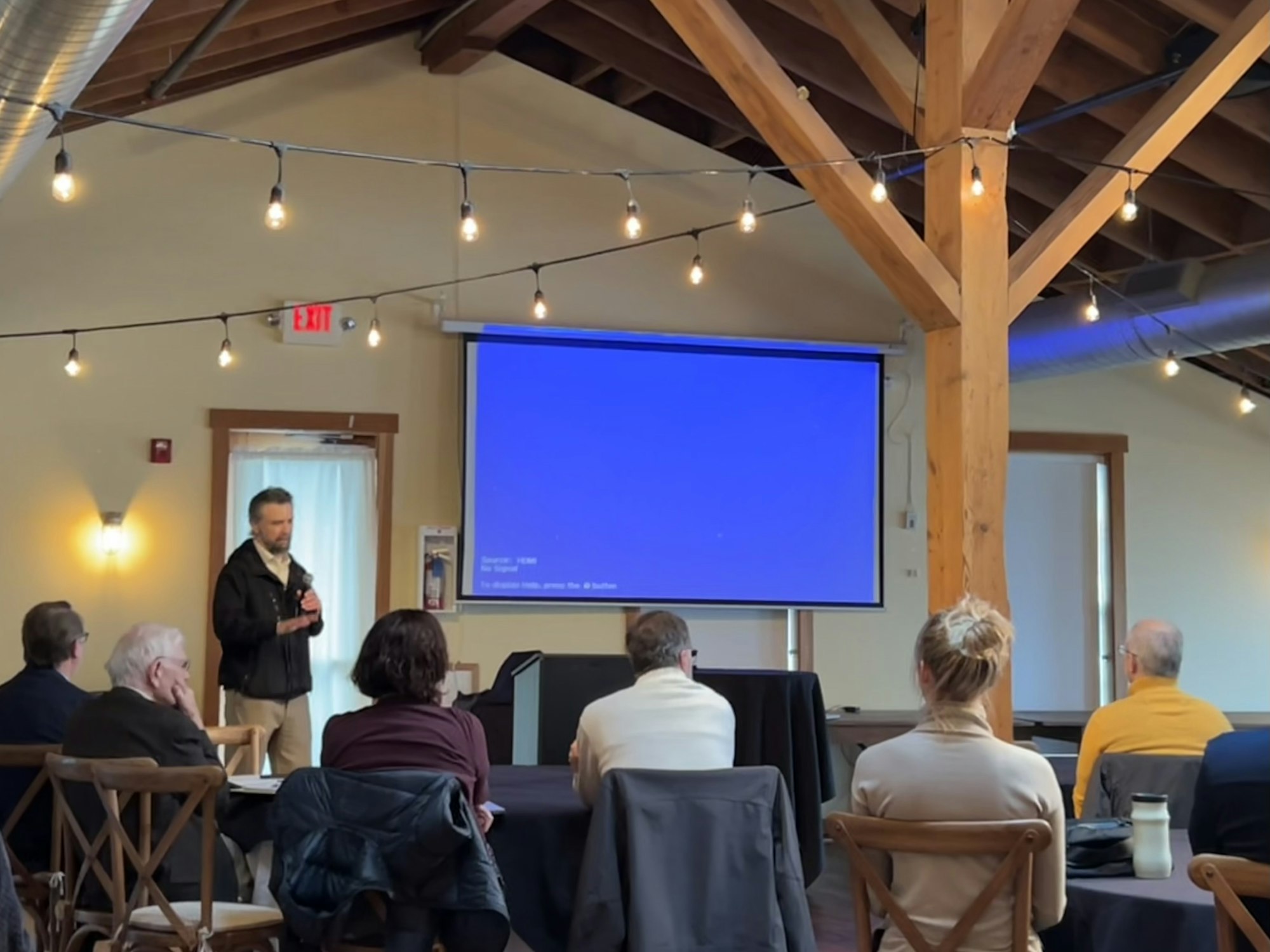 A person speaking in a room with string lights, people seated, and a blank blue projection screen.