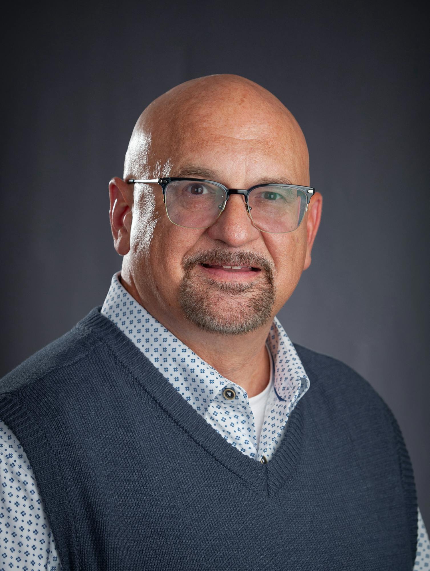 The image shows a smiling man with a bald head, glasses, wearing a dark sweater vest over a patterned shirt.