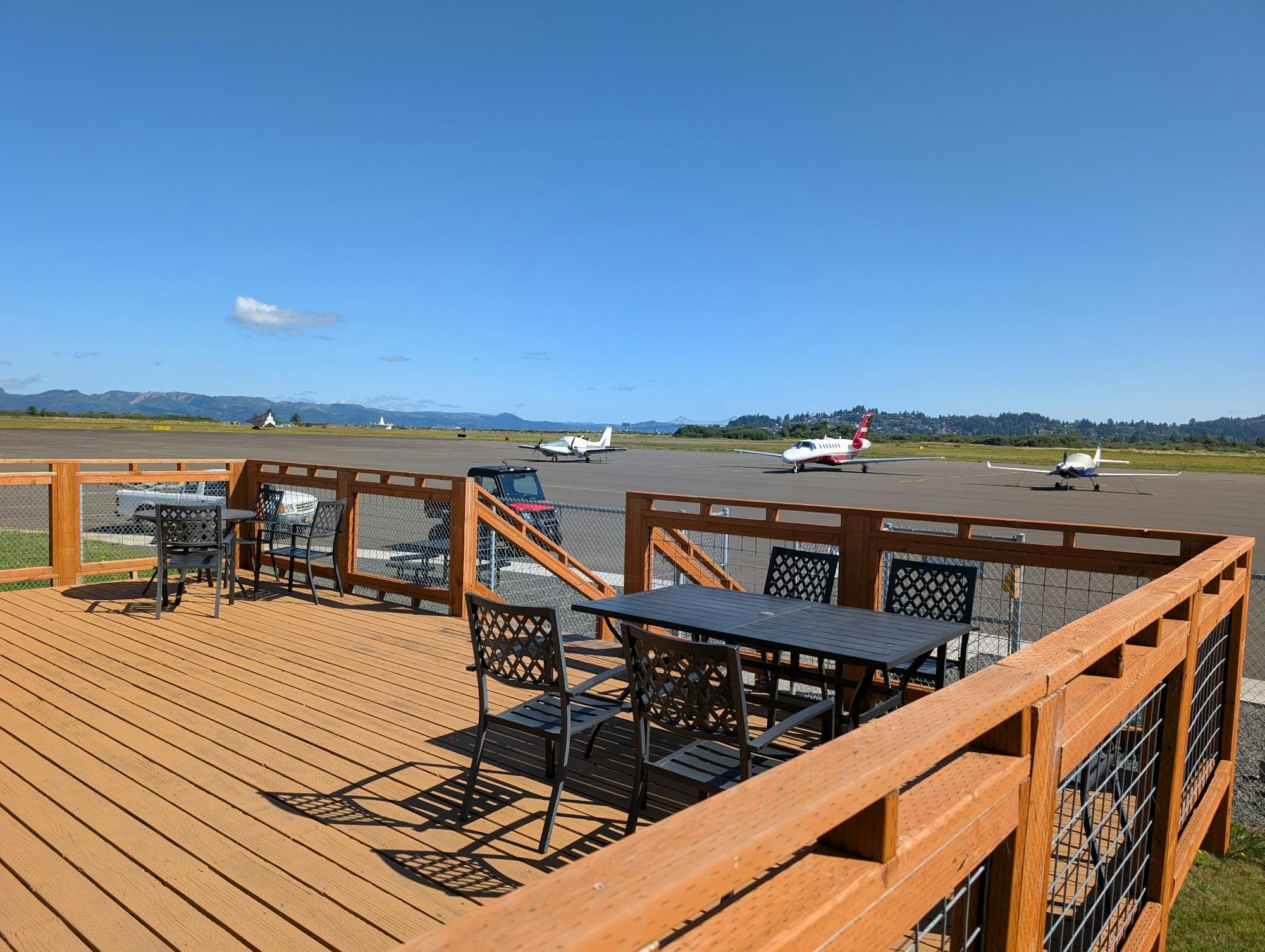 A wooden deck with tables and chairs overlooks a runway with three small airplanes. Clear sky and distant hills in the background.