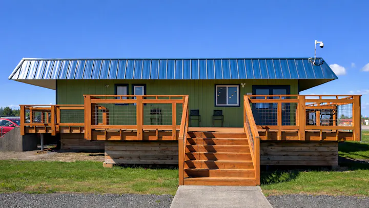 A green house with a blue metal roof, wooden deck, stairs, and chairs, set against a clear blue sky.