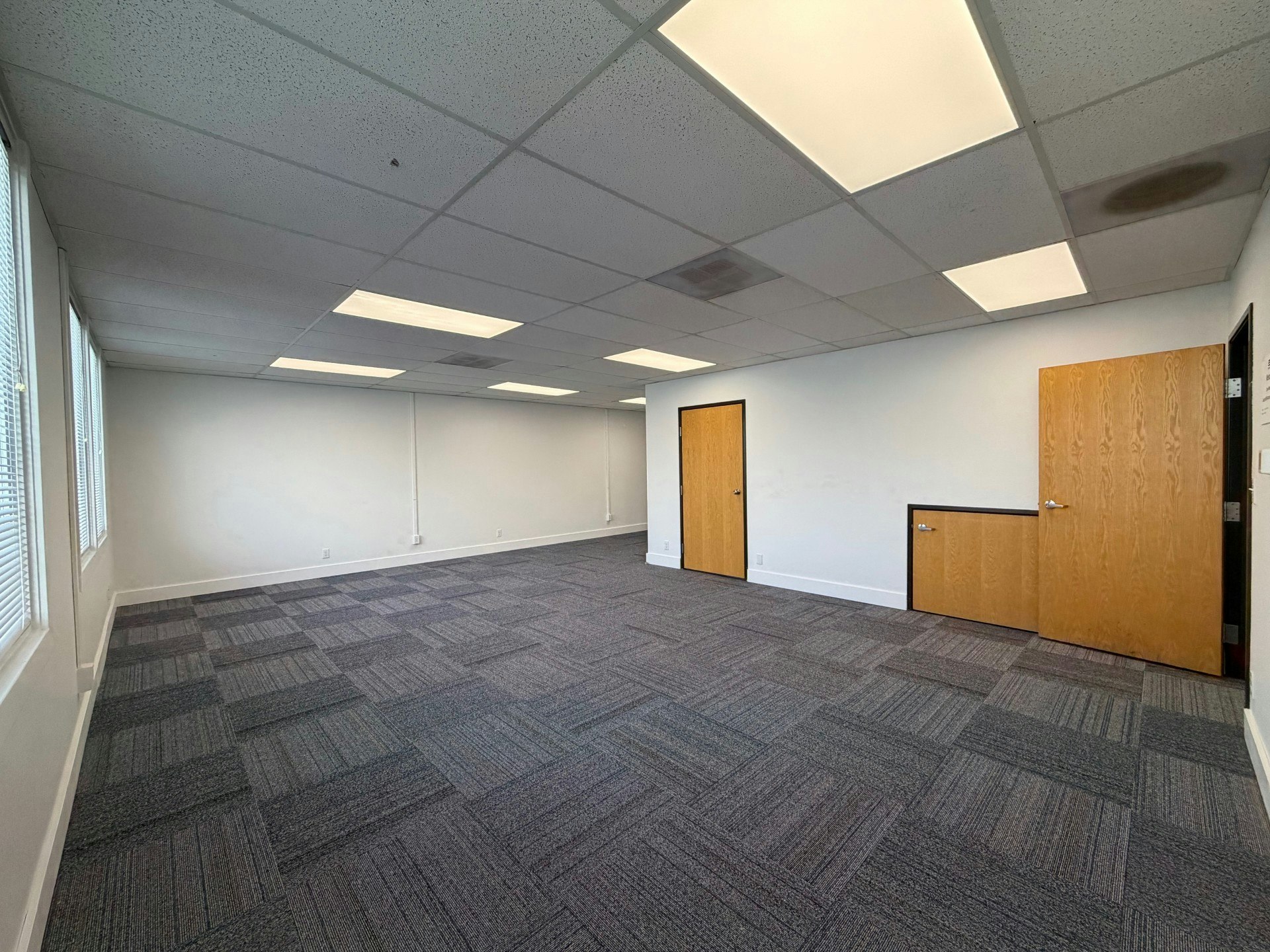 A spacious, empty office room with gray carpet, white walls, and two wooden doors. Bright overhead lighting and windows are present.