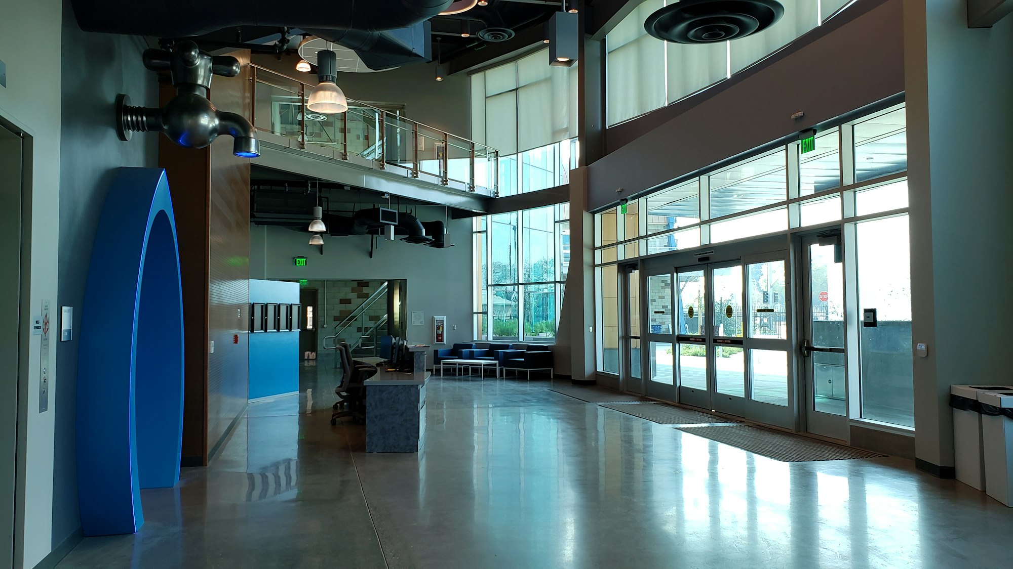 Spacious, modern lobby with high ceilings, large windows, a reception desk, and seating area. Decorative fixtures and exit signs visible.