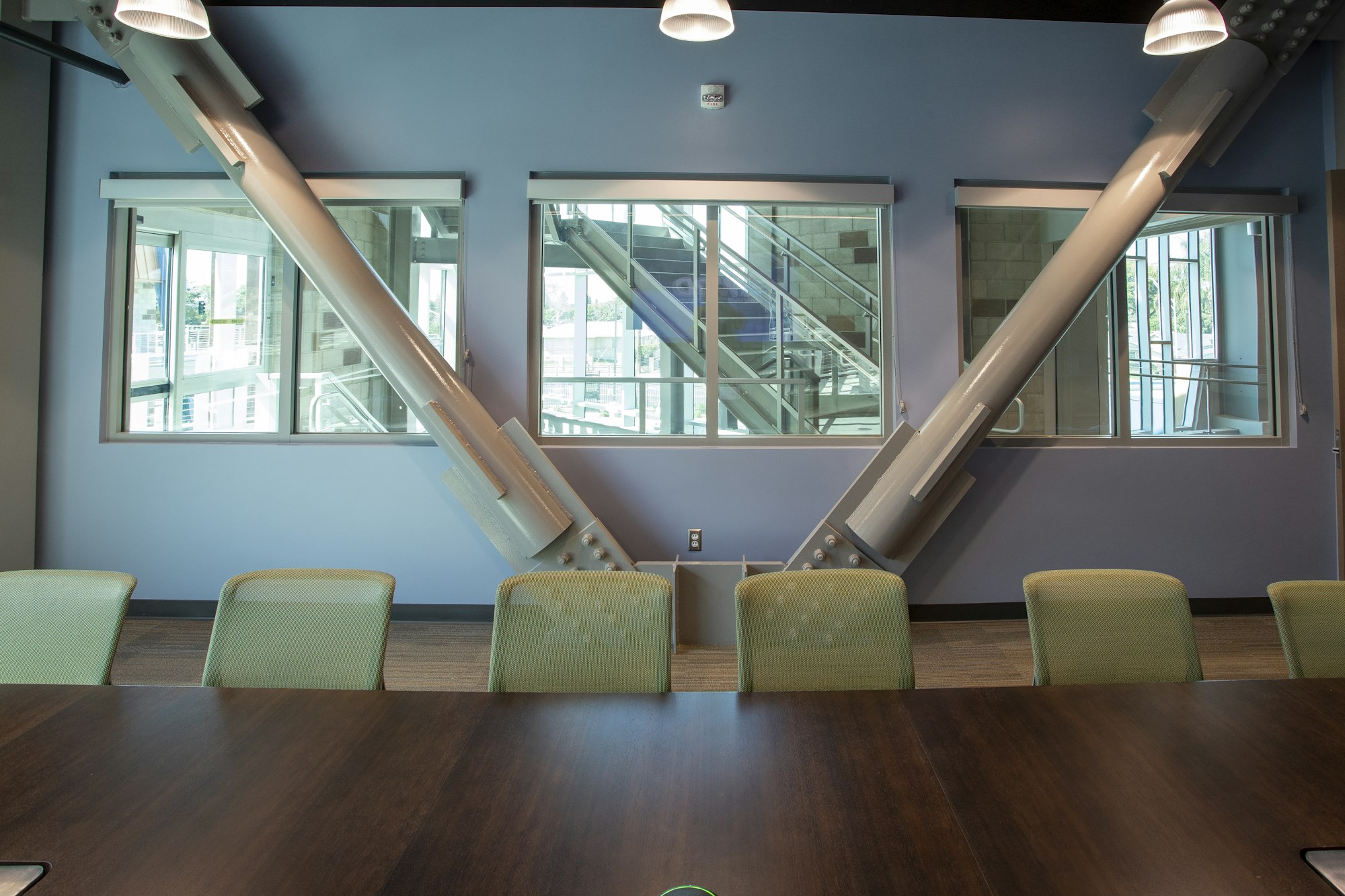 A conference room with green chairs, a wooden table, large windows, and structural beams on a blue wall.