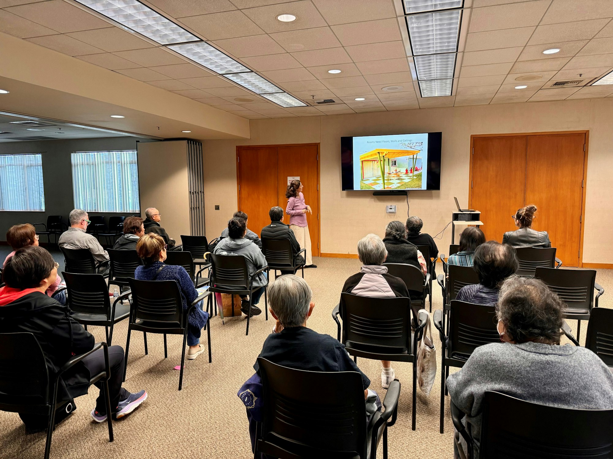 A person presents to a seated audience in a room, with a display showing an image related to a topic being discussed.