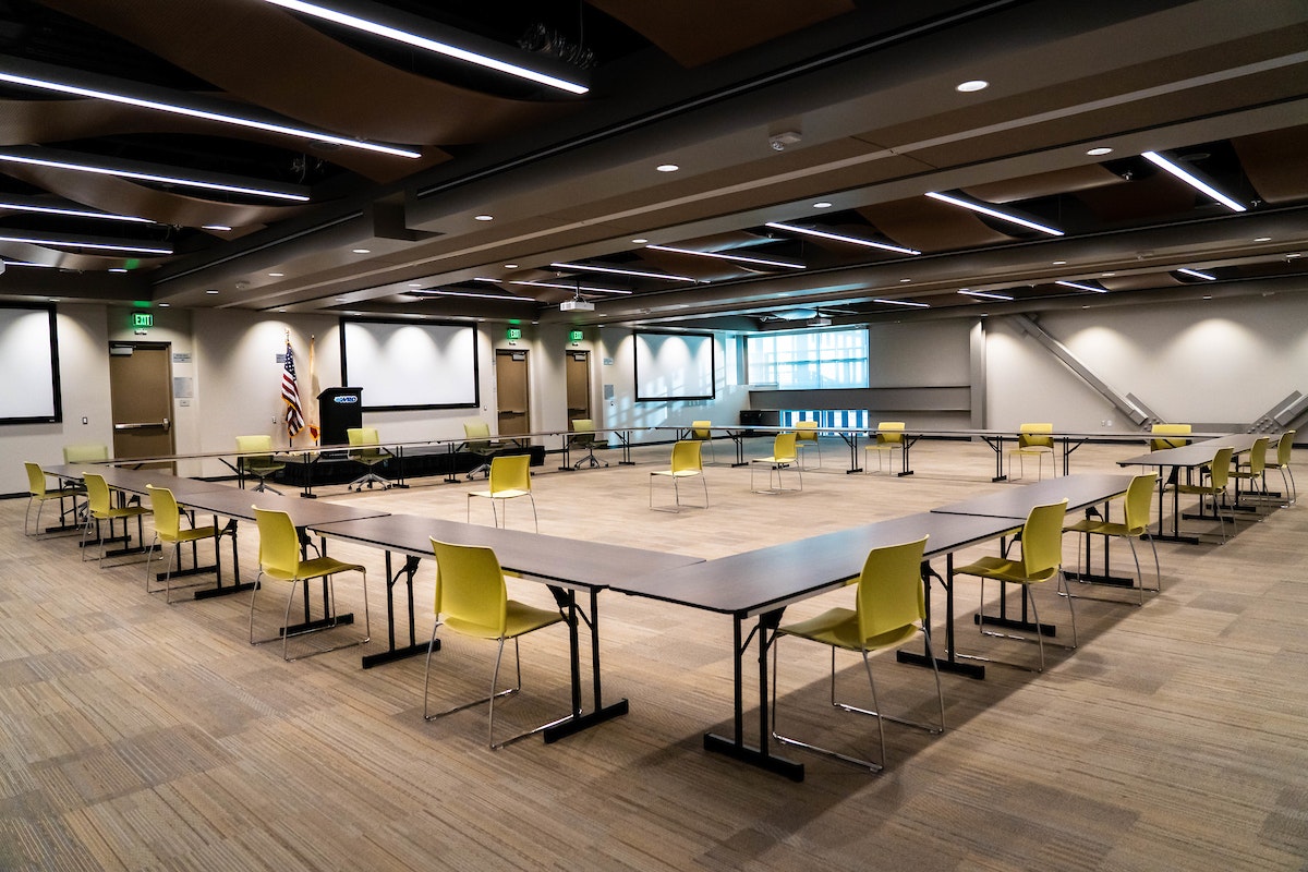 Large empty conference room with U-shaped tables and yellow chairs, ceiling lights, a projector, and an American flag.
