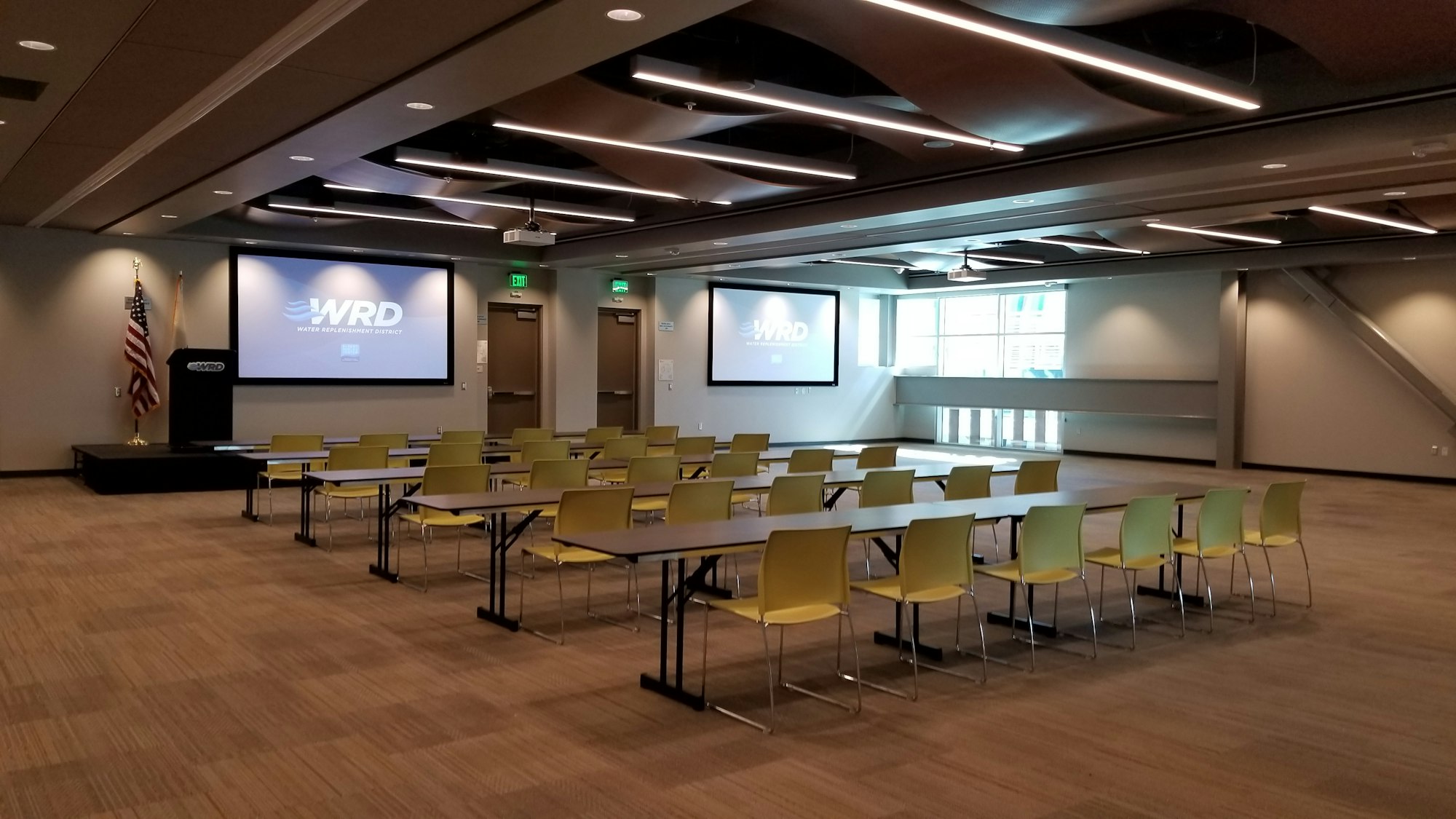 A conference room with rows of tables and chairs, two projection screens, a podium, and a flag. The room is empty.