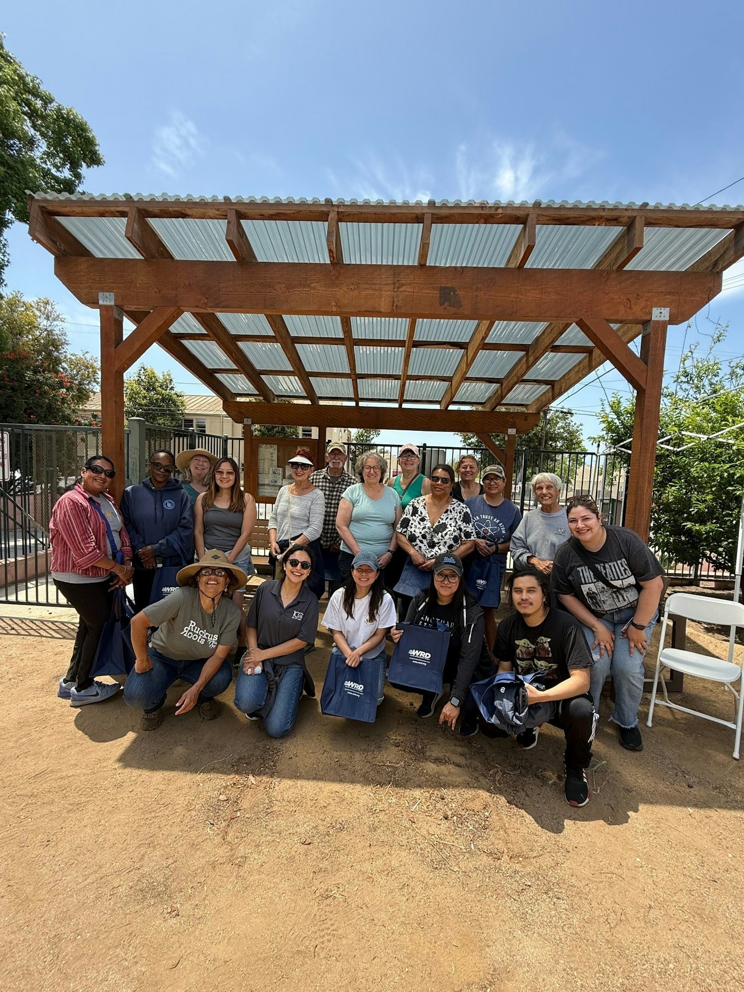 A diverse group of people poses together under a wooden structure on a sunny day, holding bags and smiling.